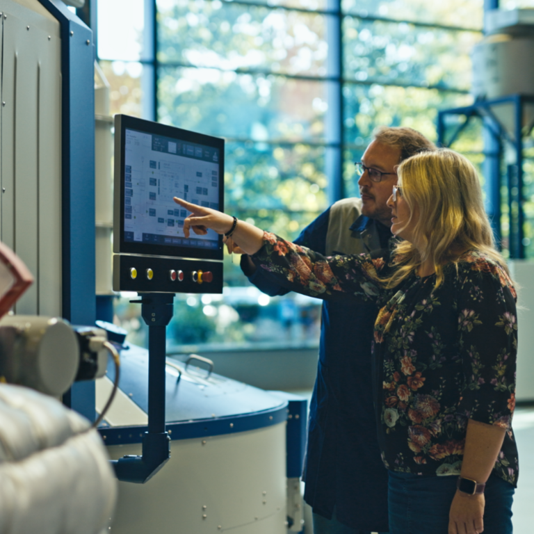 Our Technology Center offers the ideal environment for testing and training.png a man and a woman standing in front of a machine at the software control panel